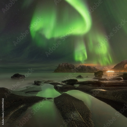 Northern lights in Lofoten islands, Norway. Green Aurora borealis. Starry sky with polar lights. Night winter landscape with aurora, sea with sky reflection, rocks, beach and snowy mountains.