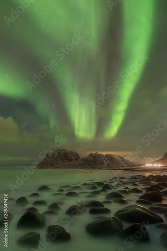 Northern lights in Lofoten islands, Norway. Green Aurora borealis. Starry sky with polar lights. Night winter landscape with aurora, sea with sky reflection, rocks, beach and snowy mountains. 