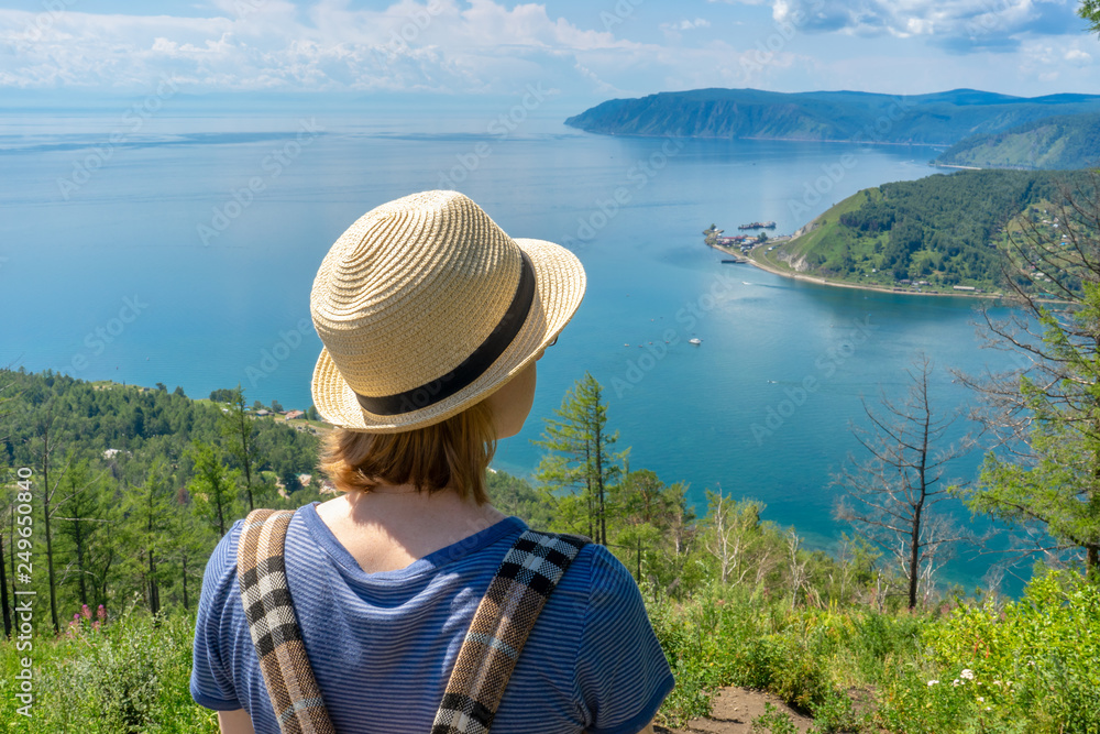 Tween tourist girl in hat and backpack standing on cliff top and admiring beautiful landscape of  Baikal lake and Angara river source. Hiking, travelling and summer vacation concept