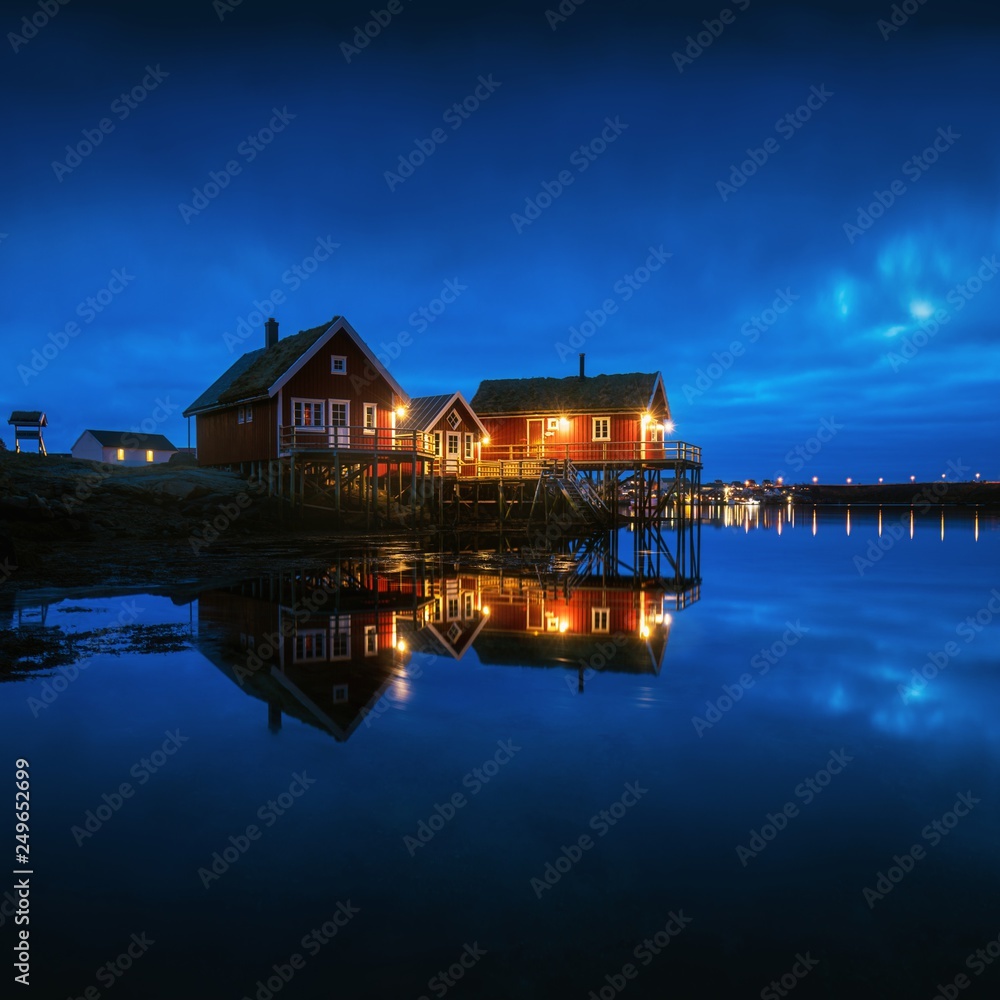 Fishing town Reine reflections in night on Lofoten islands in Norway, above the arctic circle Winter landscape with buildings, blue cloudy sky reflected in water at dusk. Norwegian rorbuer Lofoten
