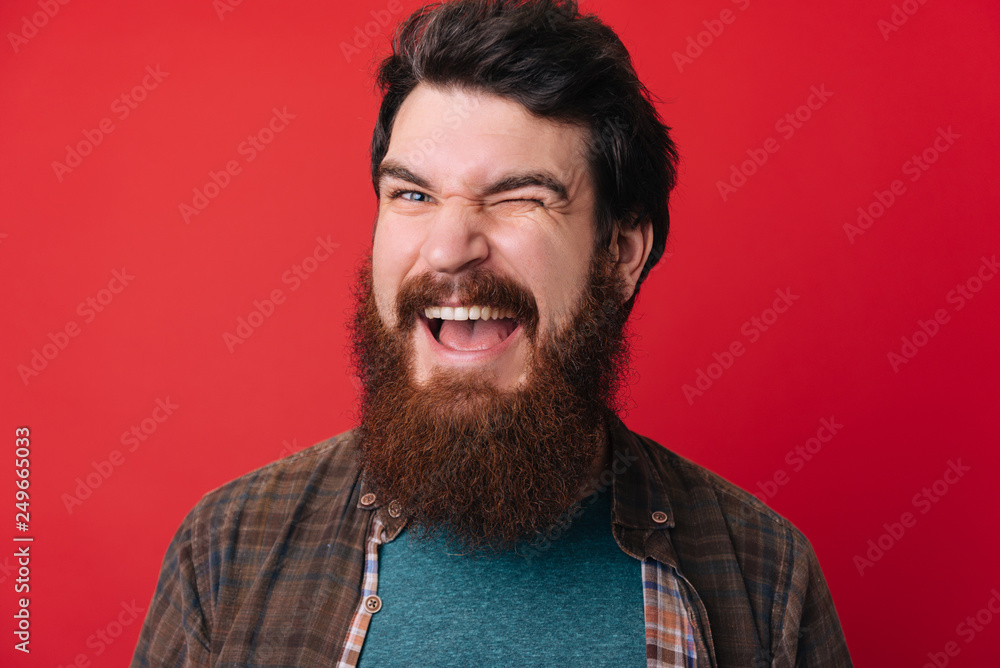 Obraz premium A handsome young bearded man doing flirting gesture with eye over red background