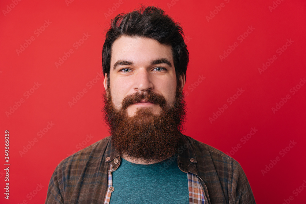 Shot of young handsome male with beard, has serious expression as listens to interlocutor, poses in studio against redbackground Shot of young handsome male with beard, has serious expression as listens to interlocutor, poses in studio against redbackground