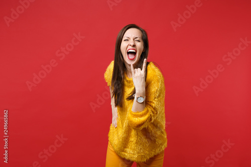 Screaming young woman in yellow fur sweater with horns up gesture, depicting heavy metal rock sign isolated on bright red background. People sincere emotions, lifestyle concept. Mock up copy space.