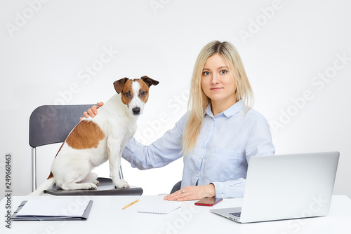 Young blonde cute caucasian woman watches straight holds her Jack Russell Terrier and sits by the desk in the office with the white background. Dog watches straight. Laptop, cell phone and documents. © Roman Tyukin