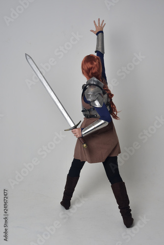 Fényképezés full length portrait of a  red haired girl wearing medieval warrior costume and steel armour, standing pose facing away from the camera on grey studio background