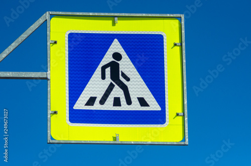 Traffic sign - a pedestrian crossing against the blue sky. A square blue-yellow crossing sign across the road. A white triangle road sign on a blue background.
