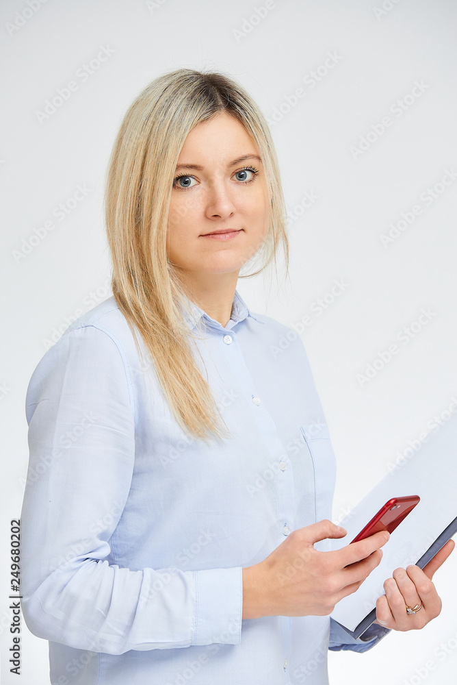 Young cute blonde caucasian woman stands sideways holding her red cell phone and document folder in the office with a white background. Young cute blonde caucasian woman stands sideways holding her red cell phone and document folder in the office with a white background.