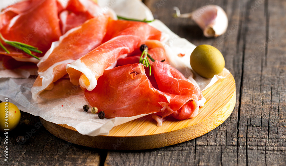 Closeup of thin slices of prosciutto with mixed olives and paprika on wooden cutting board