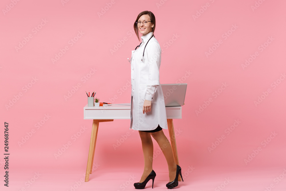 Cute female doctor stand in front of desk with pc computer, medical document in hospital isolated on pastel pink wall background. Woman in medical gown glasses stethoscope. Healthcare medicine concept Cute female doctor stand in front of desk with pc computer, medical document in hospital isolated on pastel pink wall background. Woman in medical gown glasses stethoscope. Healthcare medicine concept