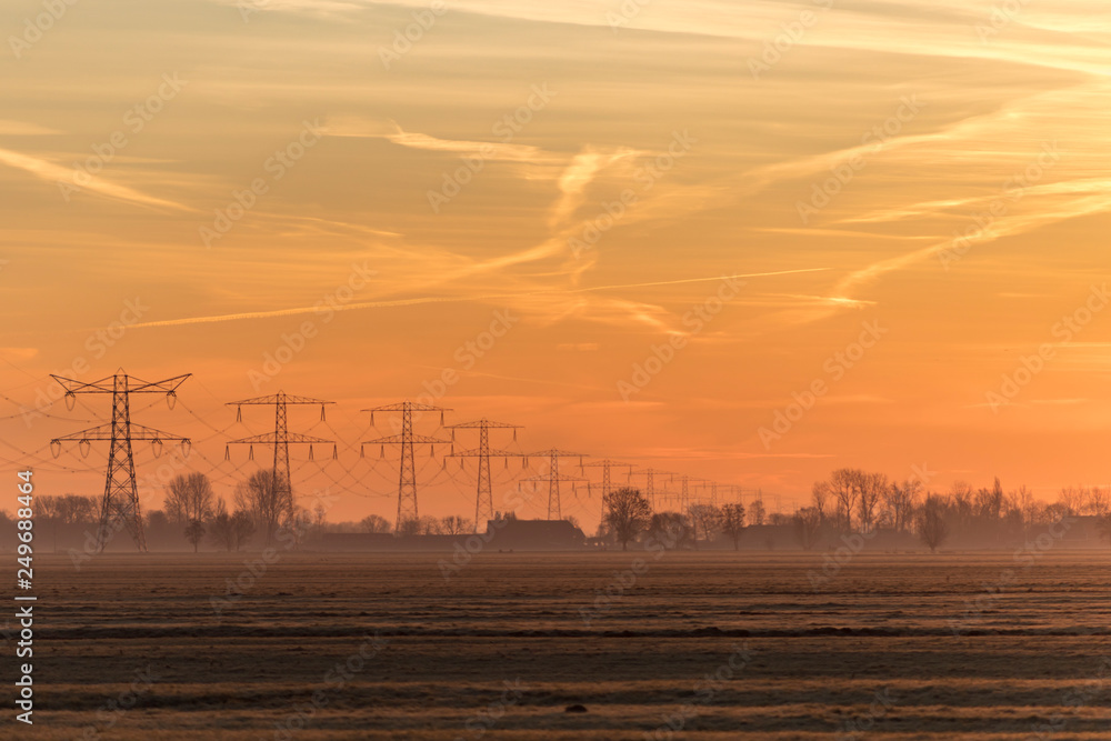 Early morning hazy rural landscape with the silhouettes of transmission towers, trees and farm houses on the background and a bright orange and yellow sky