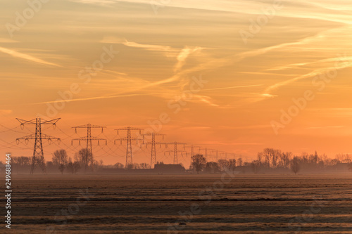 Early morning hazy rural landscape with the silhouettes of transmission towers  trees and farm houses on the background and a bright orange and yellow sky