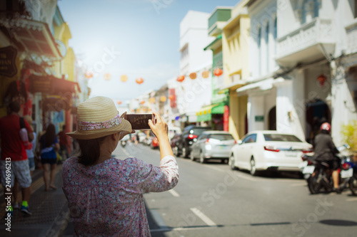 Woman traveler in casual dress taking picture in thalang road with blurred chino portuguese style building.