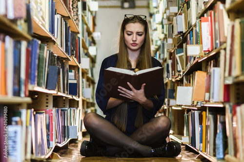 Beautiful young girl sitting among bookshelves and books on the floor in the library