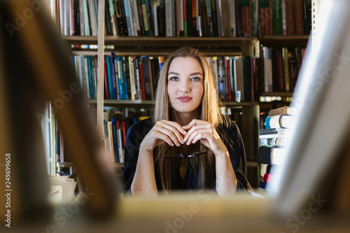 Beautiful young woman wearing glasses in the library among the bookshelves holding glasses and looking at the lens, reading, studying.