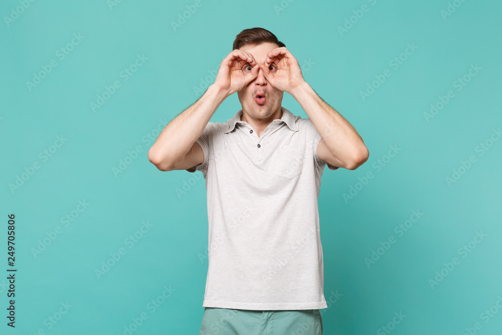 Amazed young man in casual clothes holding hands near eyes, imitating glasses or binoculars isolated on blue turquoise wall background. People sincere emotions, lifestyle concept. Mock up copy space. Amazed young man in casual clothes holding hands near eyes, imitating glasses or binoculars isolated on blue turquoise wall background. People sincere emotions, lifestyle concept. Mock up copy space.