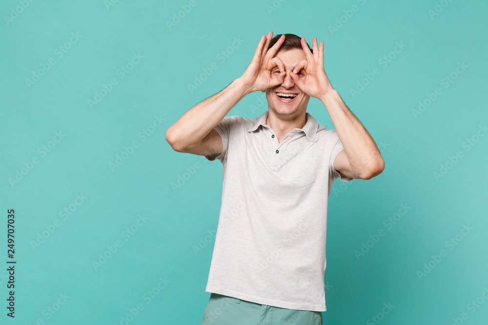 Laughing young man in casual clothes holding hands near eyes imitating glasses or binoculars isolated on blue turquoise wall background. People sincere emotions, lifestyle concept. Mock up copy space. Laughing young man in casual clothes holding hands near eyes imitating glasses or binoculars isolated on blue turquoise wall background. People sincere emotions, lifestyle concept. Mock up copy space.