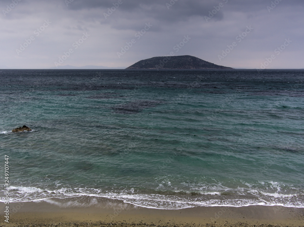 Small island, sea, clouded sky, and sand. Turquoise colored water and small wave foam on sandy beach with small island on background. Small island, sea, clouded sky, and sand. Turquoise colored water and small wave foam on sandy beach with small island on background.