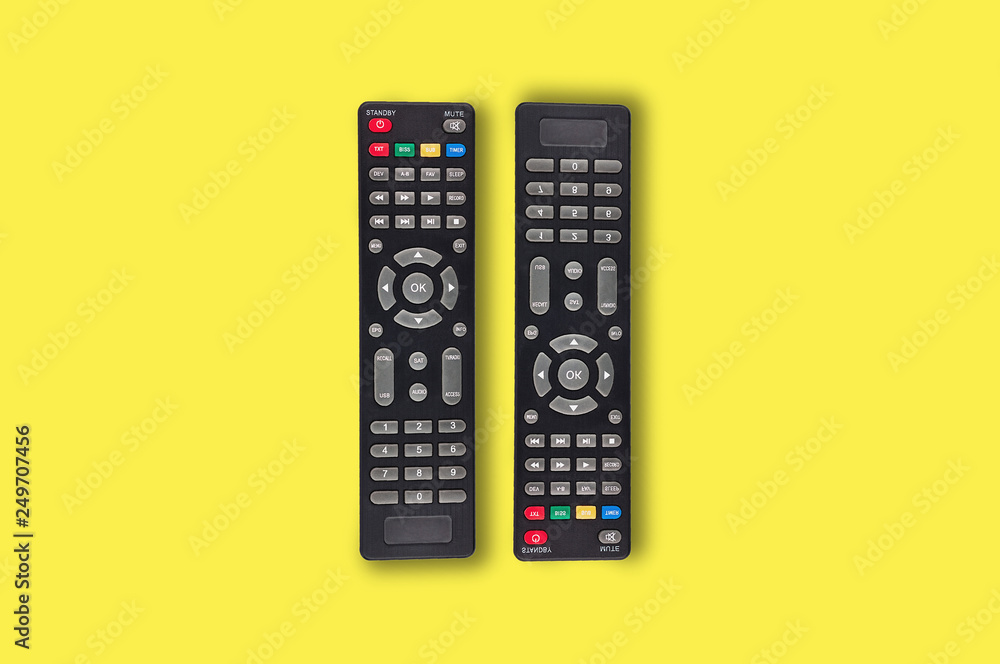 Pair of black plastic remote controls for different multimedia devices in center on yellow background. Top view Pair of black plastic remote controls for different multimedia devices in center on yellow background. Top view