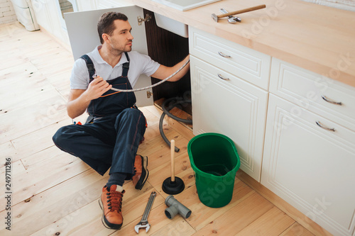 Young handyman sit on floor in kitchen. He put new hose under sink. Guy concentrated. Daylight. Bucket with tools on floor.