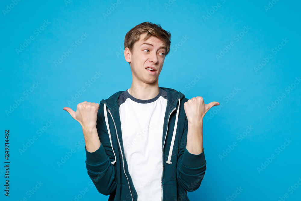 Portrait of bewildered confident young man in casual clothes standing, pointing thumbs aside isolated on blue wall background in studio. People sincere emotions, lifestyle concept. Mock up copy space. Portrait of bewildered confident young man in casual clothes standing, pointing thumbs aside isolated on blue wall background in studio. People sincere emotions, lifestyle concept. Mock up copy space.