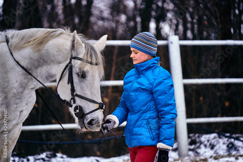 Young pretty woman in blue jacket and sports hat on a walk with a white horse on a winter cloudy day. A horse licks its hands to its owner.