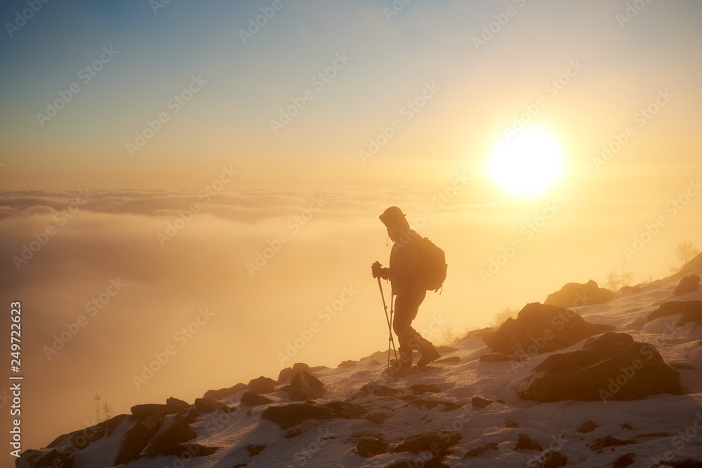 Tourist hiker with backpack and trekking poles climbing on rocky snowy mountain steep slope on background of foggy valley filled with white puffy clouds, raising sun and blue sky at dawn.