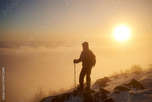 Back view of tourist hiker with backpack and hiking sticks resting on rocky mountain peak on copy space background of beautiful foggy valley filled with white puffy clouds at dawn at winter.