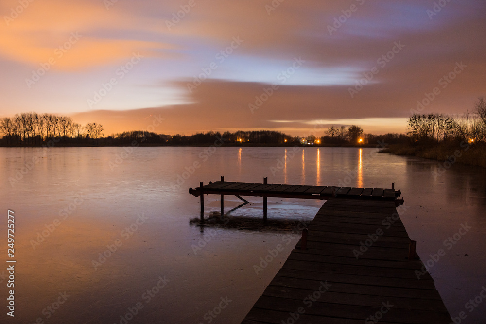 Wooden bridge with planks on a frozen lake and clouds after sunset