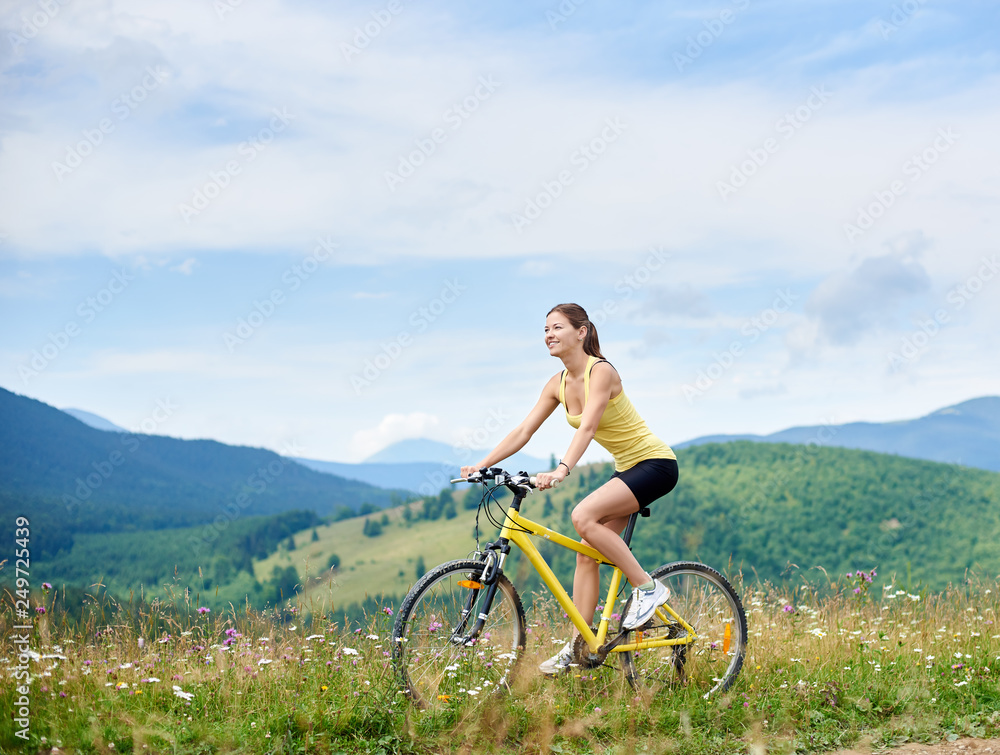 Attractive happy girl cyclist riding on yellow mountain bike on a grassy hill, enjoying summer day in the mountains. Outdoor sport activity, lifestyle concept Attractive happy girl cyclist riding on yellow mountain bike on a grassy hill, enjoying summer day in the mountains. Outdoor sport activity, lifestyle concept