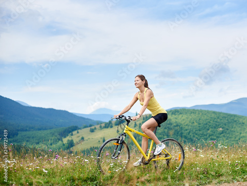 Attractive happy girl cyclist riding on yellow mountain bike on a grassy hill, enjoying summer day in the mountains. Outdoor sport activity, lifestyle concept