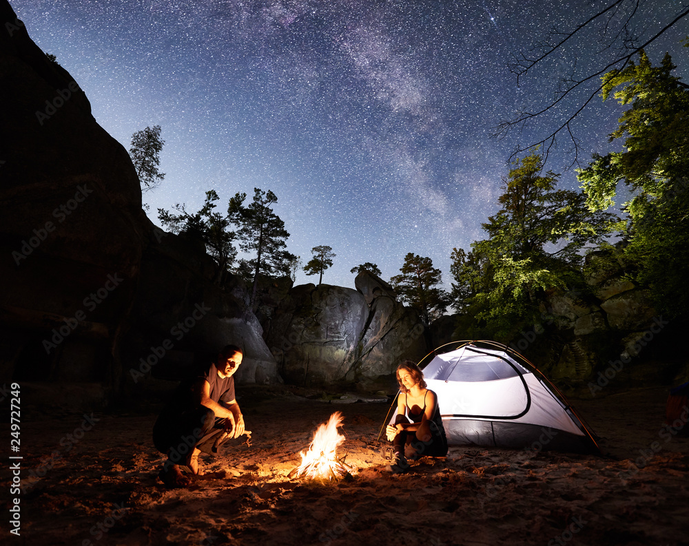 Young couple travellers man and woman resting beside camp, campfire and glowing tourist tent at night. On background beautiful night starry sky full of stars and Milky way, mountain rocks, trees - obrazy, fototapety, plakaty 