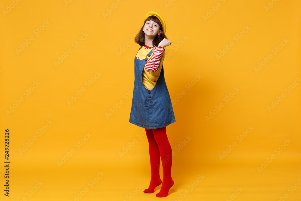 Full length portrait of pretty girl teenager in french beret, denim sundress holding hair isolated on yellow wall background in studio. People sincere emotions, lifestyle concept. Mock up copy space. Full length portrait of pretty girl teenager in french beret, denim sundress holding hair isolated on yellow wall background in studio. People sincere emotions, lifestyle concept. Mock up copy space.