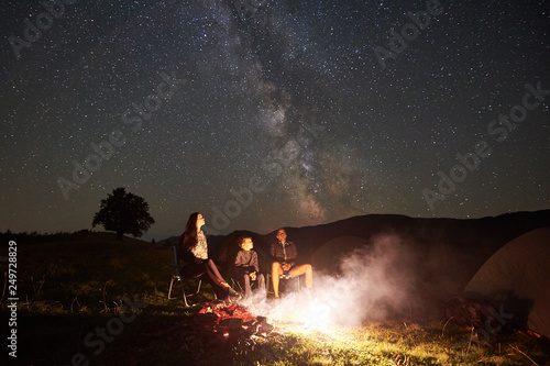 Young tourist family having a rest together in mountains, sitting on chairs beside camp, campfire and tent at summer night. Mother, father and son enjoying night starry sky full of stars and Milky way