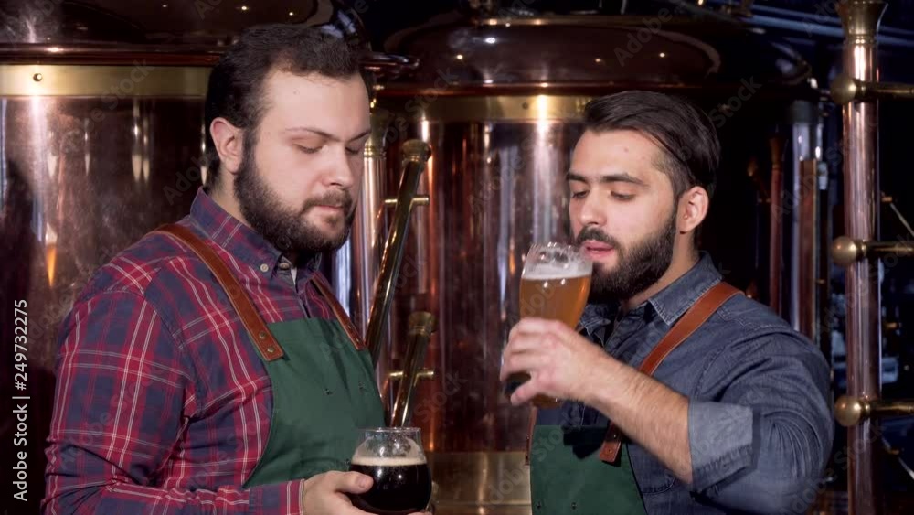 Two professional brewers clinking glasses drinking delicious craft beer together. Handsome bearded brewer celebrating with his colleague, enjoying freshly brewed beer Two professional brewers clinking glasses drinking delicious craft beer together. Handsome bearded brewer celebrating with his colleague, enjoying freshly brewed beer