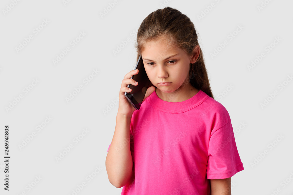 Sad small girl talking on modern mobile phone in a studio. Unhappy little schoolgirl heard bad news from her relatives by smartphone. Gloomy girl looking down uses mobile phone to communicate Sad small girl talking on modern mobile phone in a studio. Unhappy little schoolgirl heard bad news from her relatives by smartphone. Gloomy girl looking down uses mobile phone to communicate