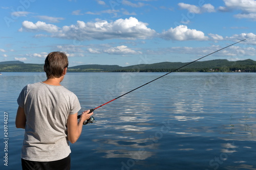 Woman is fishing from a beautiful blue lake against a mountainous shore and the sky with clouds. Bright shot.