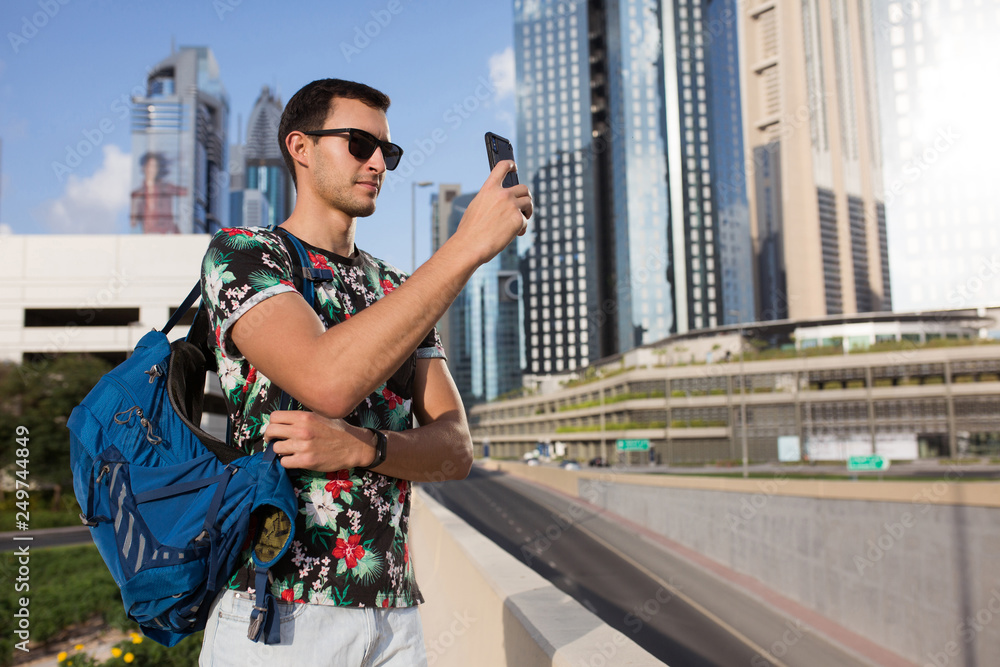 Young attractive man in t-shirt and sunglasses with backpack dreamily taking photos on cellphone with beautiful city skyscrapers on background outdoor