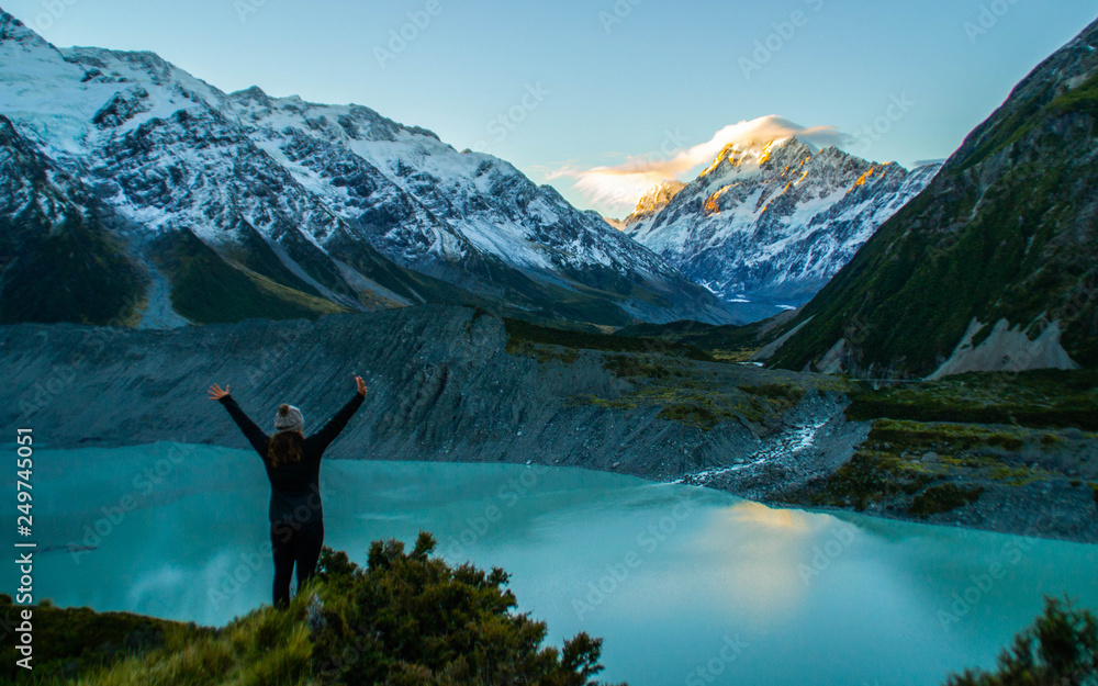 Travel New Zealand. Young tourist/backpacker woman standing in front of the highest New Zealand mountain having hands up in the air. Happiness or winner outdoor background concept. Travel New Zealand. Young tourist/backpacker woman standing in front of the highest New Zealand mountain having hands up in the air. Happiness or winner outdoor background concept.