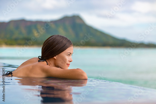 Luxury vacation travel woman relaxing in infinity swimming pool on summer beach resort. Asian girl tourist on wellness spa relaxation watching sunset outside in nature landscape background. © Maridav
