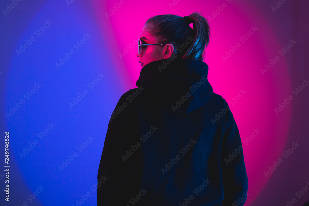 Portrait of fashion young girl in a black sweater with a hood and round sunglasses in red and blue neon light in the studio - obrazy, fototapety, plakaty 