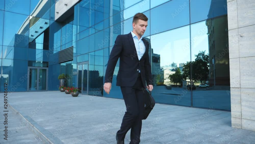 Young businessman in suit walking along city street near modern building. Confident guywith briefcase goes on office background. Handsome man on his way from office. Slow motion Young businessman in suit walking along city street near modern building. Confident guywith briefcase goes on office background. Handsome man on his way from office. Slow motion
