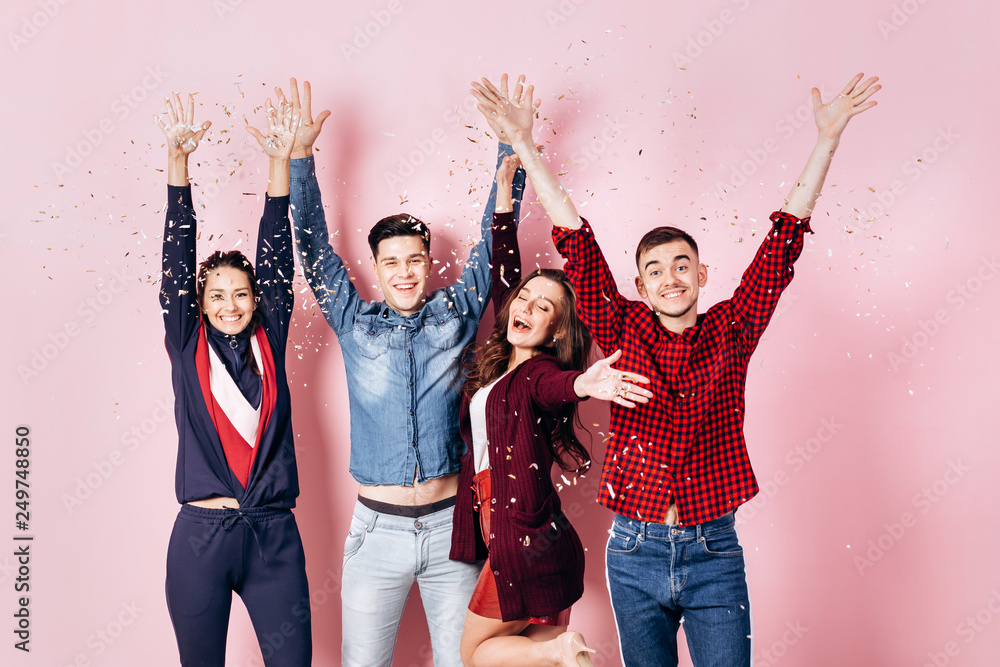 Cheerful company of two girls and two guys dressed in stylish clothes are standing and having fun with confetti on a pink background in the studio