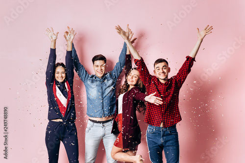 Cheerful company of two girls and two guys dressed in stylish clothes are standing and having fun with confetti on a pink background in the studio