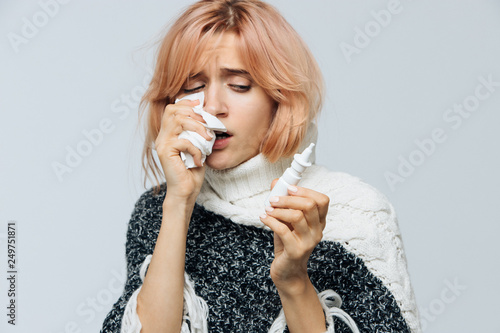 Cute unhealthy Caucasian woman with paper napkin sneezing, using nasal spray to help herself, experiences allergy symptoms, caught a cold. Rhinitis, cold, sickness, allergy concept