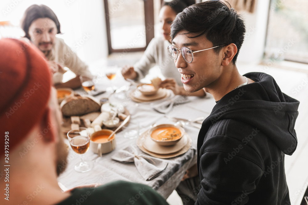 Young smiling asian man with dark hair in eyeglasses and hoodie sitting at the table happily looking aside. Group of international friends having lunch spending time together in cozy cafe - obrazy, fototapety, plakaty 