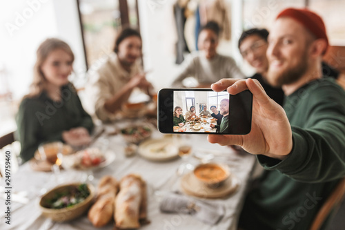 Group of attractive international friends sitting at the table with food spending time together. Young colleagues joyfully taking photo on modern cellphone having lunch in cafe