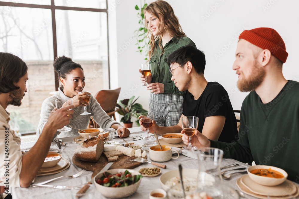 Pretty woman with wavy hair standing near table happily with colleagues. Group of young  international friends dreamily holding glasses of wine in hands having lunch together in cozy cafe