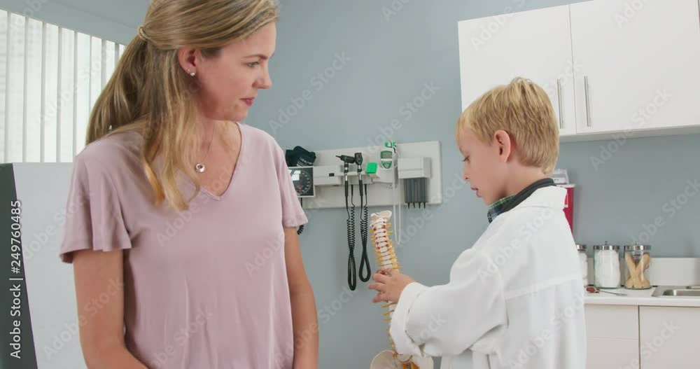 Cute child pretending to be a doctor with his mother while in pediatricians office showing her a model of the human spine. Little boy wearing oversized lab coat in exam room. Slow motion 4k Cute child pretending to be a doctor with his mother while in pediatricians office showing her a model of the human spine. Little boy wearing oversized lab coat in exam room. Slow motion 4k