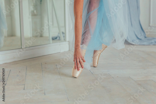 Hands of ballerina in blue tutu skirt puts on pointe shoes on leg in white light hall. Young classical ballet dancer woman in dance class