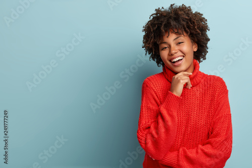 Carefree young woman with dark skin, keeps hand under chin, laughs happily, shows white teeth, wears oversized red sweater, isolated over blue background with empty space for your information